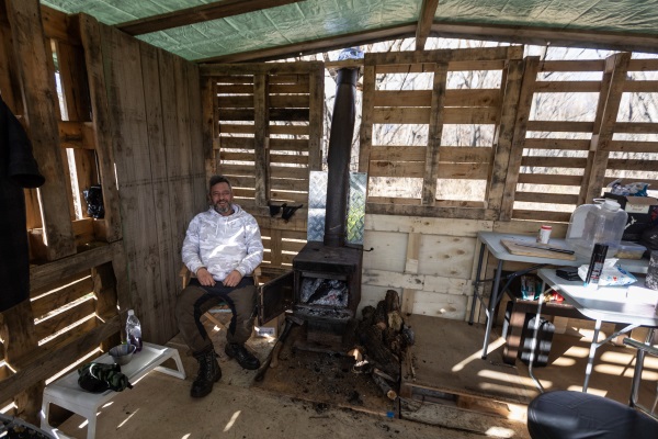 Jeremiah Greenbank warms himself next to a log burner in a shack he built from pallets on a Doc...